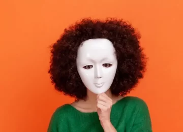 Unknown woman with Afro hairstyle wearing green casual style sweater hiding her face behind white mask, hiding her personality, anonymous. Indoor studio shot isolated on orange background.