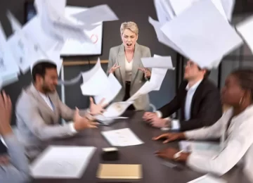 a senior businesswoman screaming in anger during a meeting in an office with paperwork falling around.